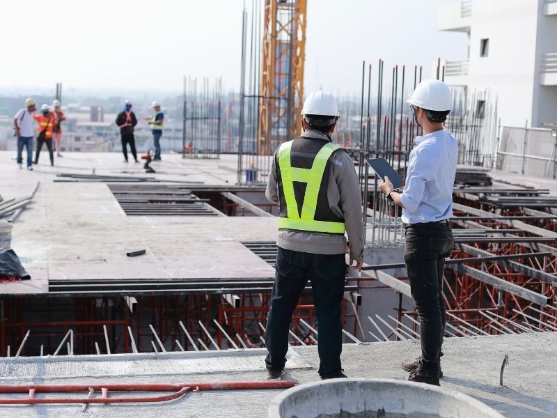 Construction site with workers in hard hats; two supervisors discuss on a ledge, one with a tablet to review plans.