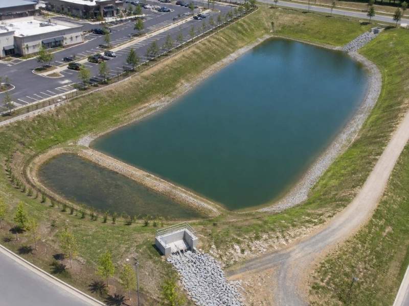 Aerial view of a detention pond beside a commercial development in Raleigh, North Carolina, showing stormwater storage, outlet structure, maintenance access and landscaped pond slopes.